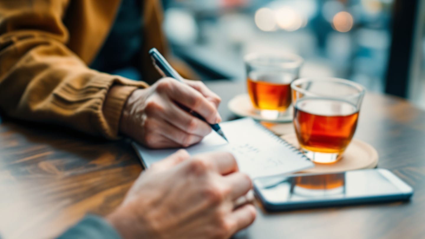 Kafe masasında not defterine yazan el, yanında yüzükoyun telefon ve çay